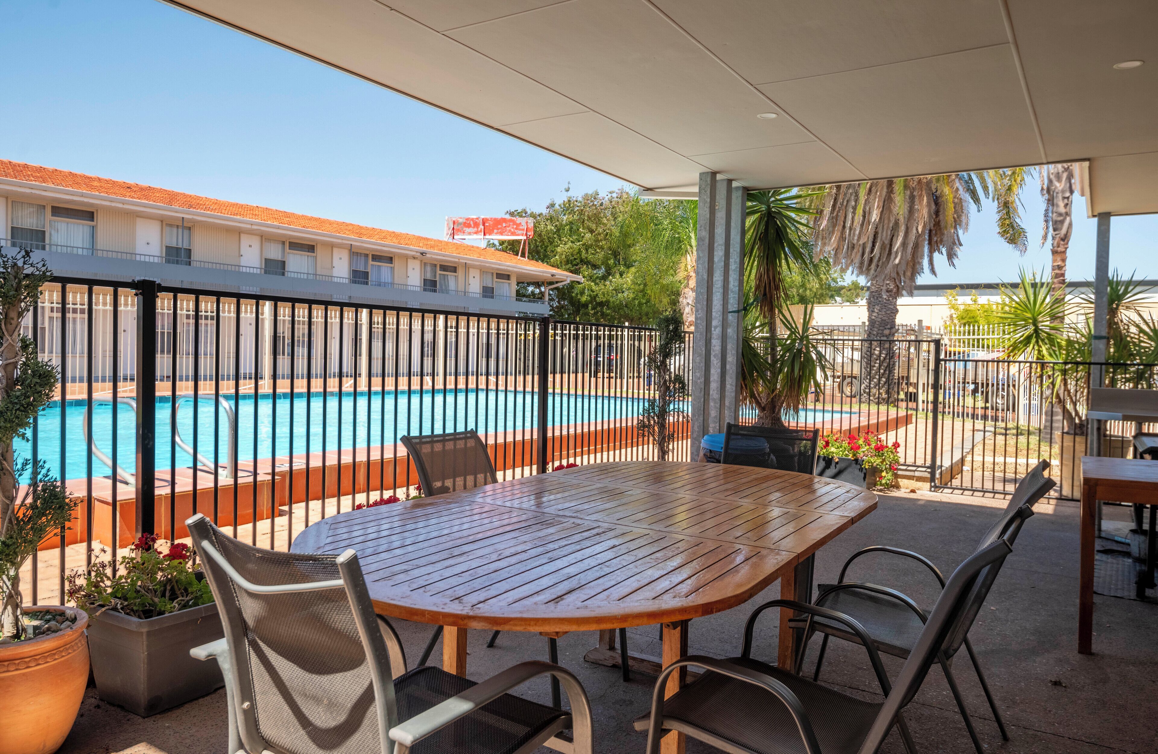 Un espace aménagé au bord de la piscine, avec une table et des chaises en bois, entouré d’une clôture en métal, et un bâtiment avec un toit rouge en arrière-plan.