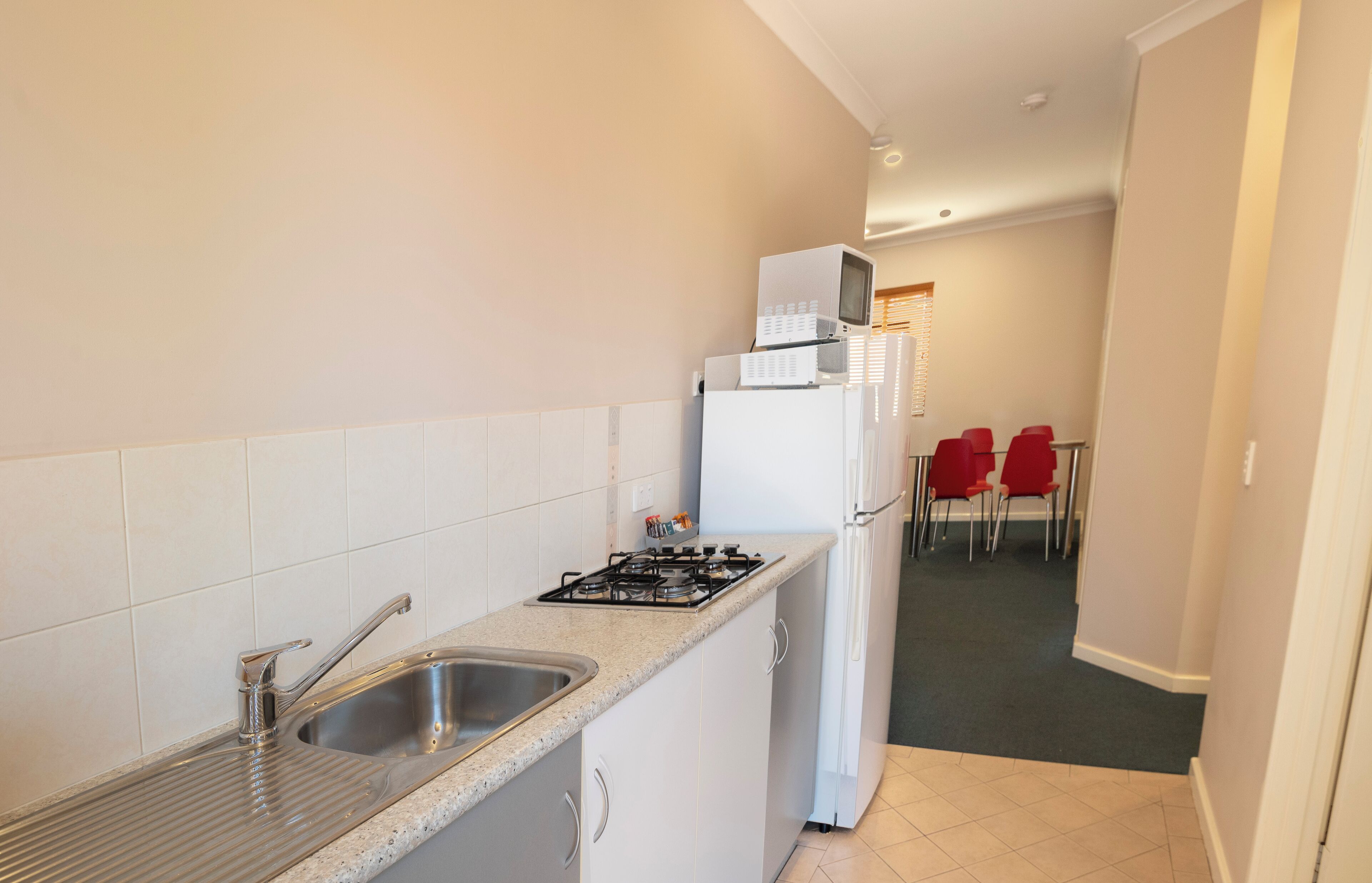 A compact kitchen with a sink, stove, and microwave. There are red chairs in the adjacent dining area.