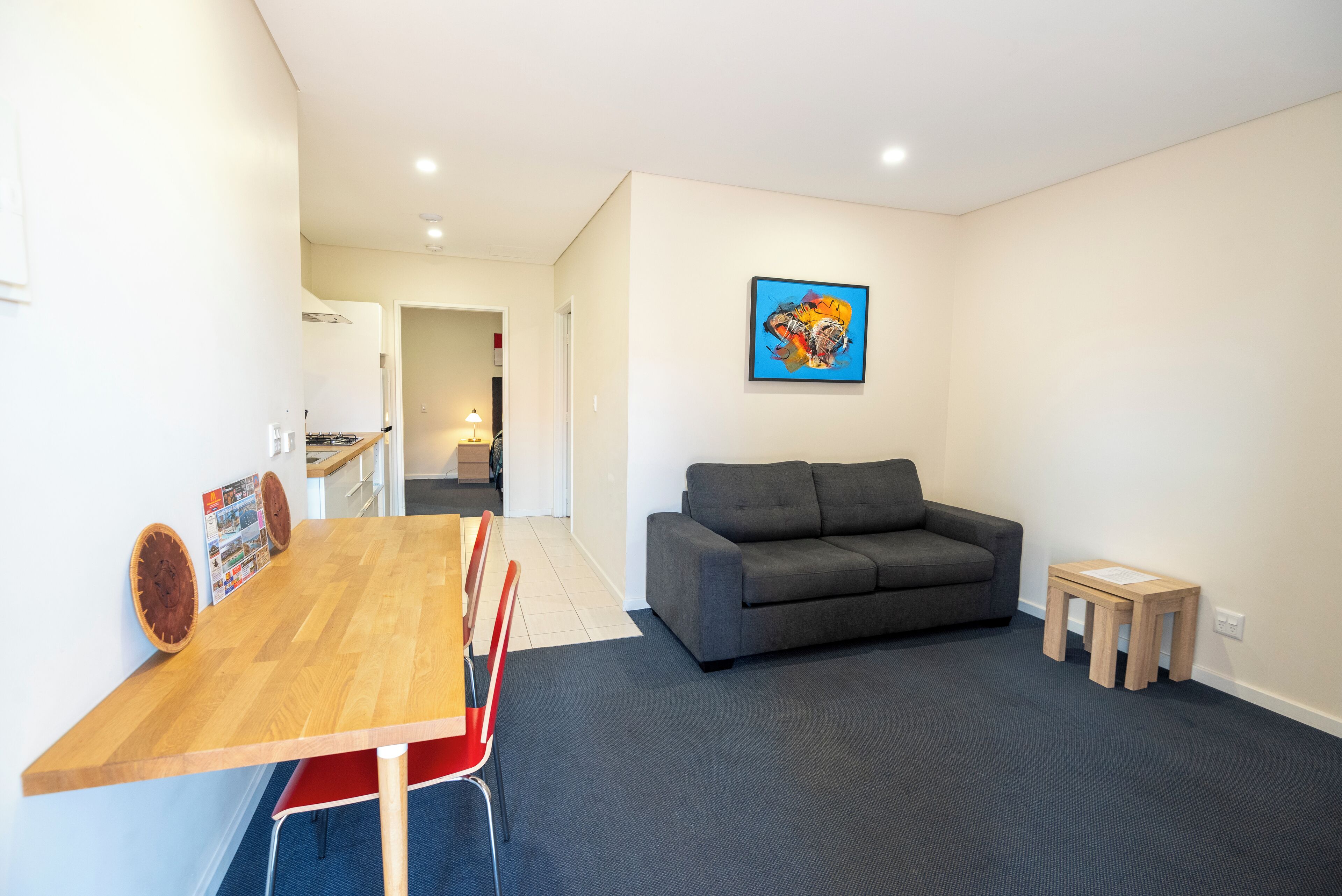 A modern living room with a grey sofa, a wooden dining table with red chairs, and a small wooden stool.