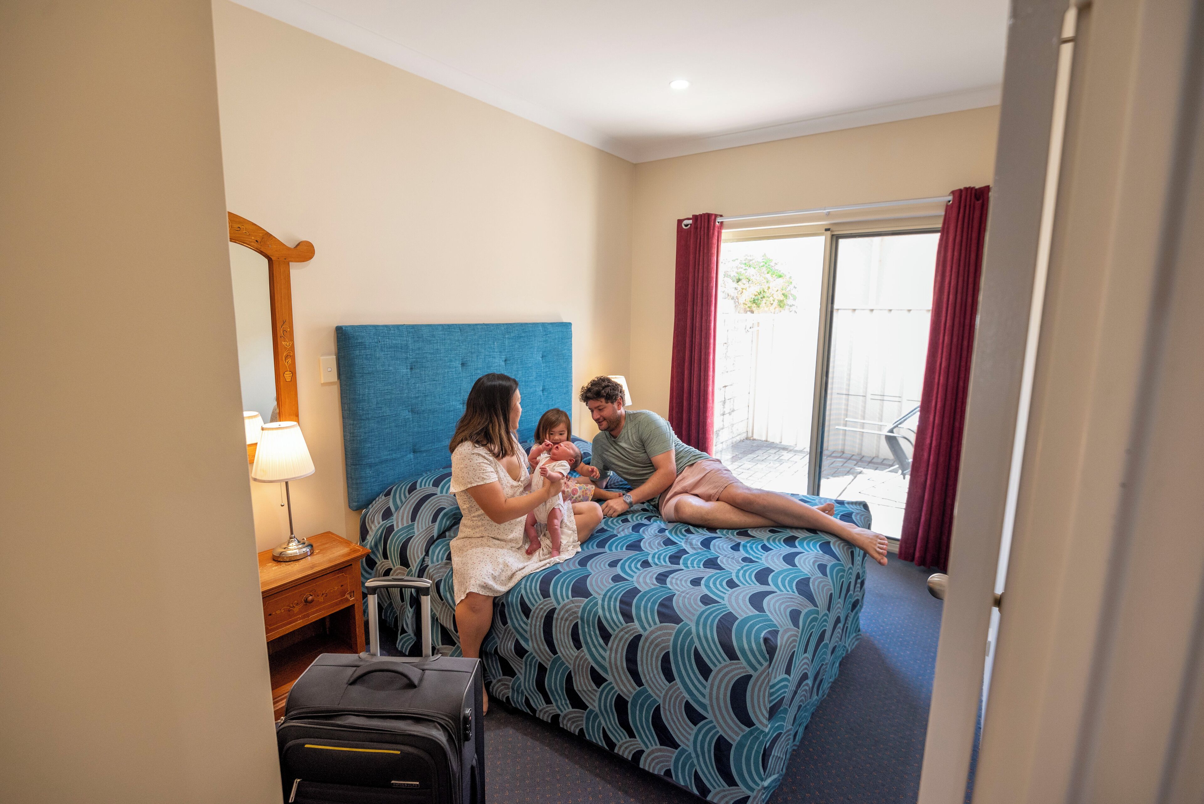 A hotel room with a bed, a nightstand with a lamp, a suitcase, and a window with curtains.