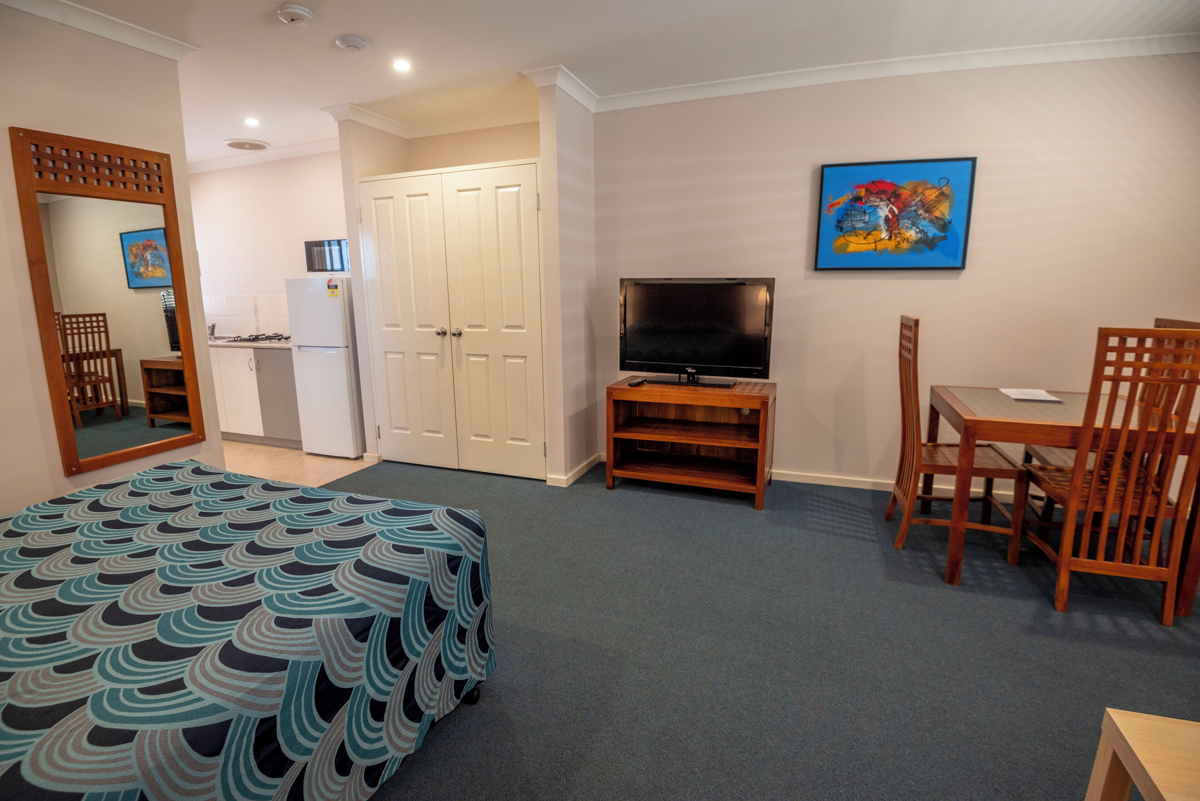 A hotel room with a bed, a TV, a desk, and a chair. There is a wardrobe and a small kitchenette with a fridge and a stove.