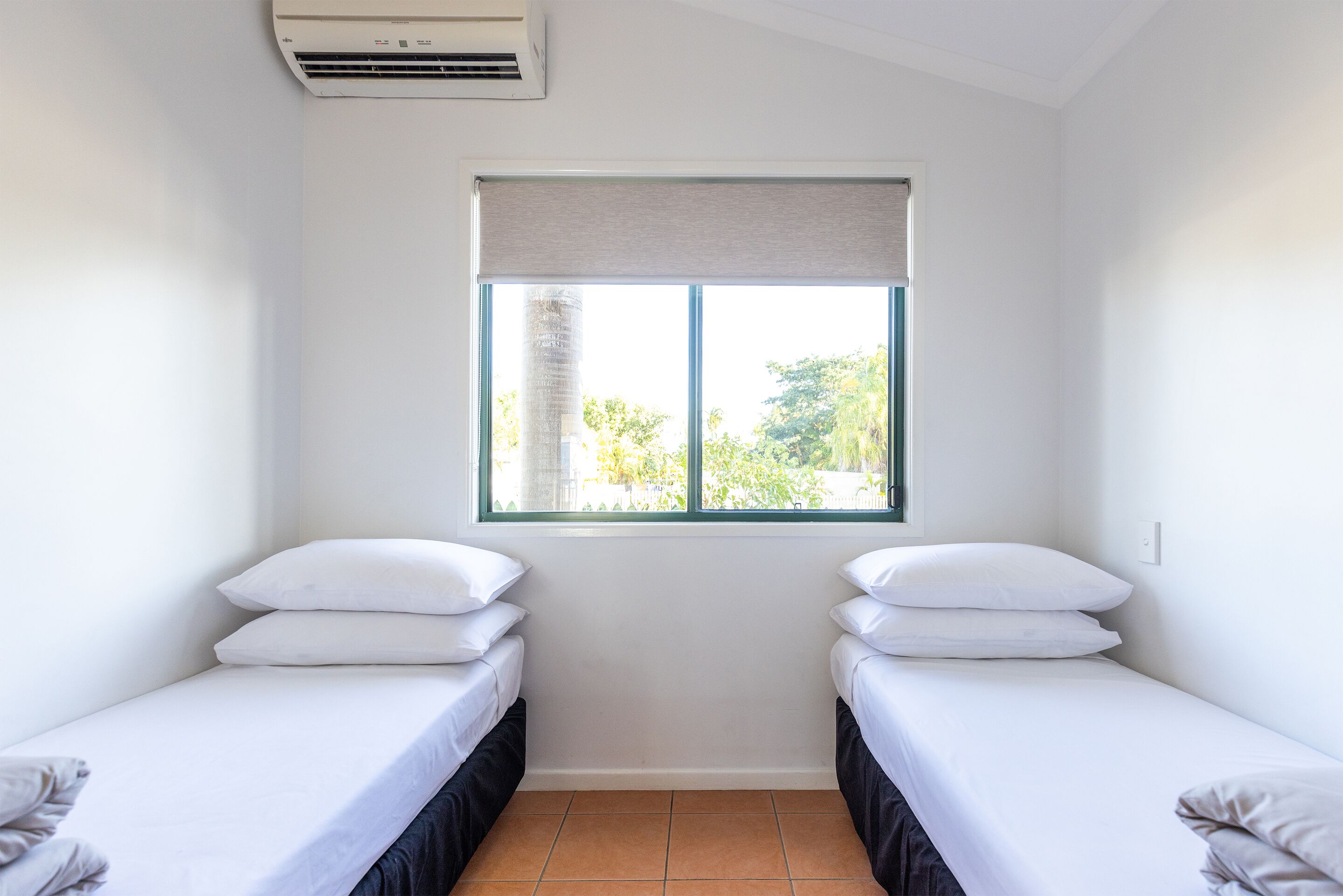 A room with two single beds, a window with a view of greenery, and an air conditioning unit.