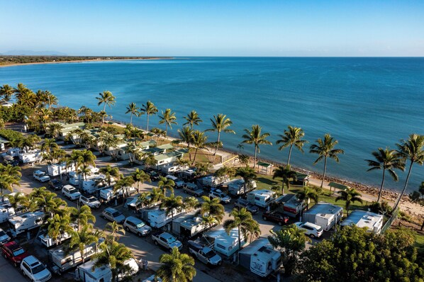 Un camping con vehículos recreativos y automóviles estacionados cerca de una playa con palmeras y el océano.