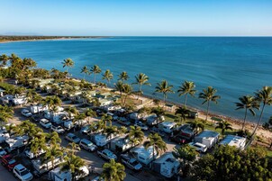 Un camping con vehículos recreativos y automóviles estacionados cerca de una playa con palmeras y el océano.