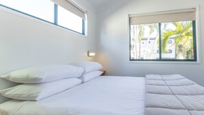 Een net opgemaakt bed met witte lakens en kussens, een nachtkastje met een lamp, en een raam met uitzicht op palmbomen en een gebouw.