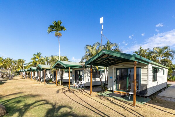 A row of small, single-story cabins with green roofs and open sides, surrounded by palm trees under a clear blue sky. - NRMA Bowen Beachfront Holiday Park (Bowen)