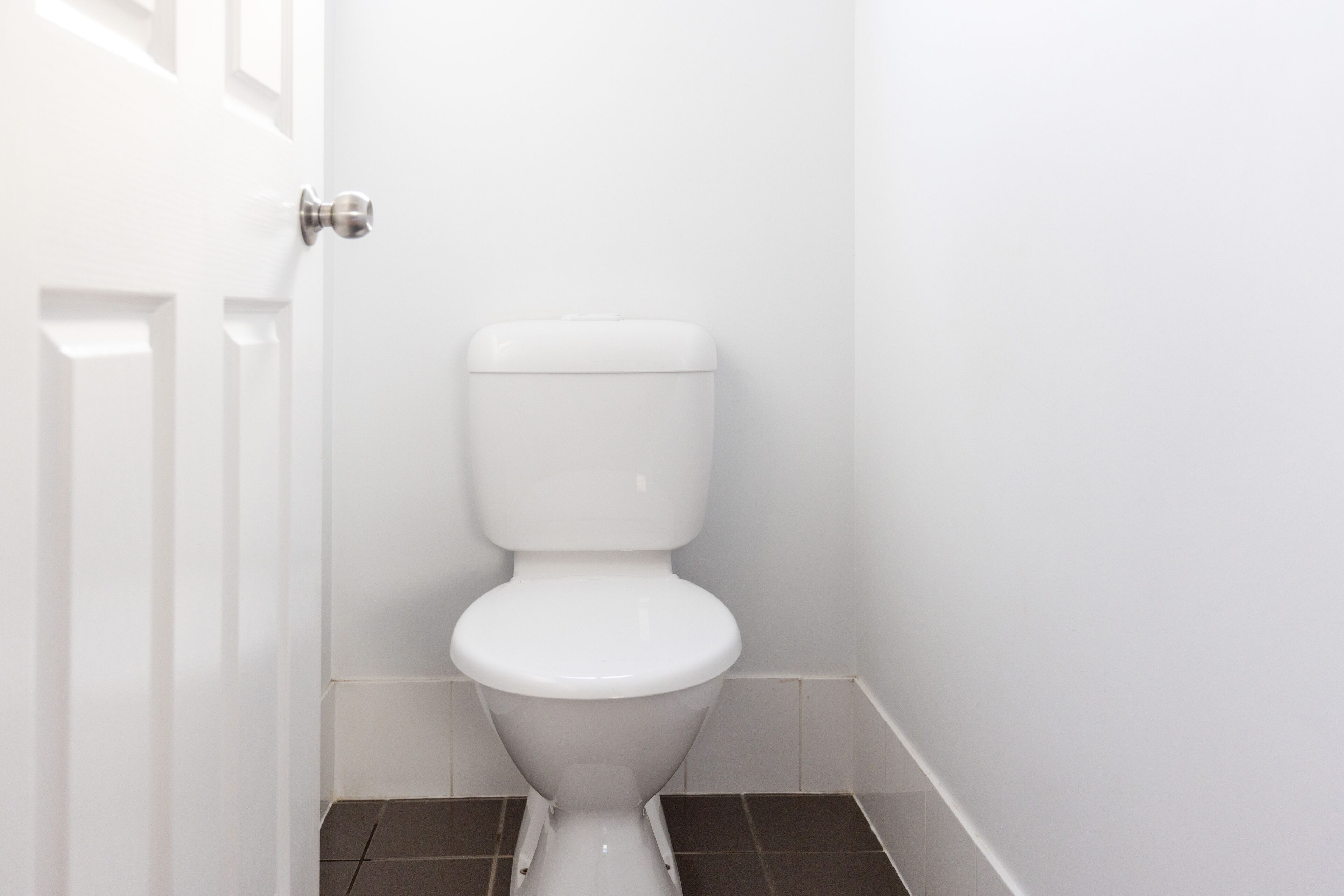 Un inodoro blanco en un baño con una puerta blanca y suelo de baldosas oscuras.