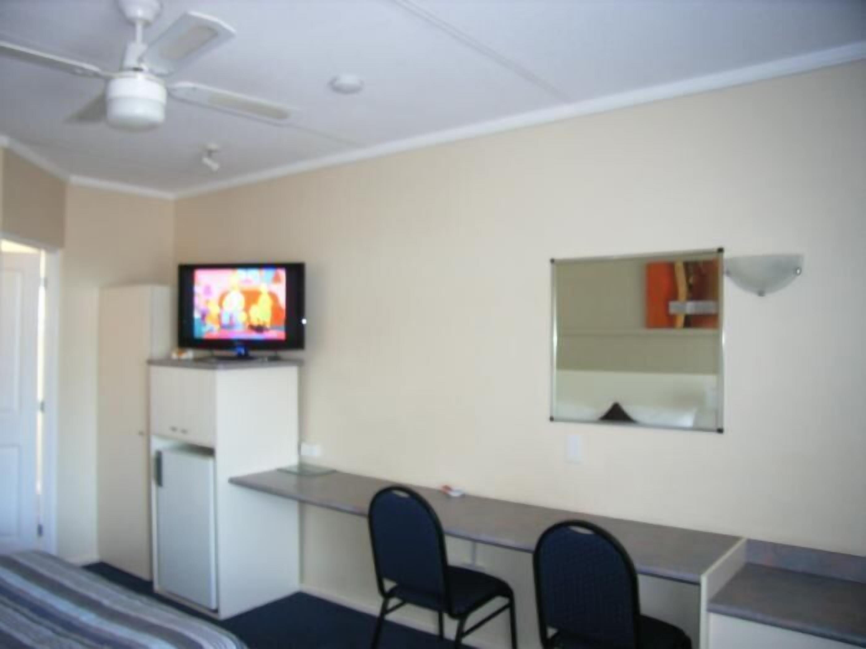 A hotel room with a bed, a desk with two chairs, a television, a ceiling fan, and a mirror.