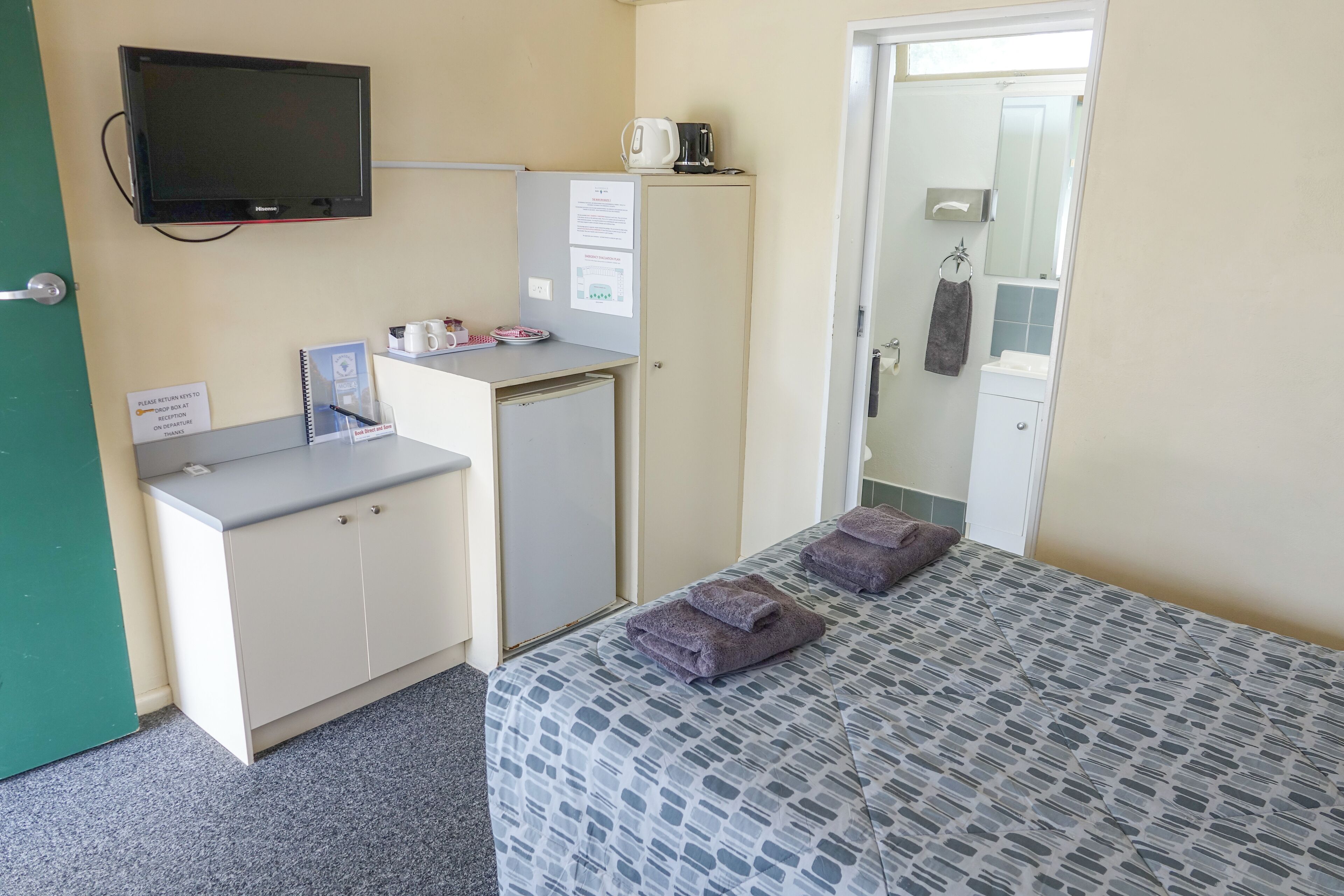 Quarto de hotel com uma cama, uma escrivaninha com computador, um mini frigorífico, uma televisão e um banheiro visível por uma porta aberta.