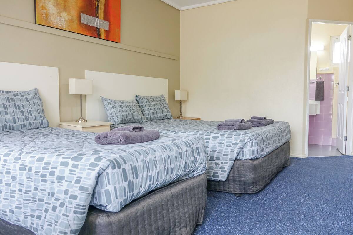 Une chambre d’hôtel avec deux lits, chacun recouvert d’une couvre-lit à motifs et agrémenté d’oreillers, une table de chevet avec une lampe, et une salle de bain visible à travers une porte ouverte.