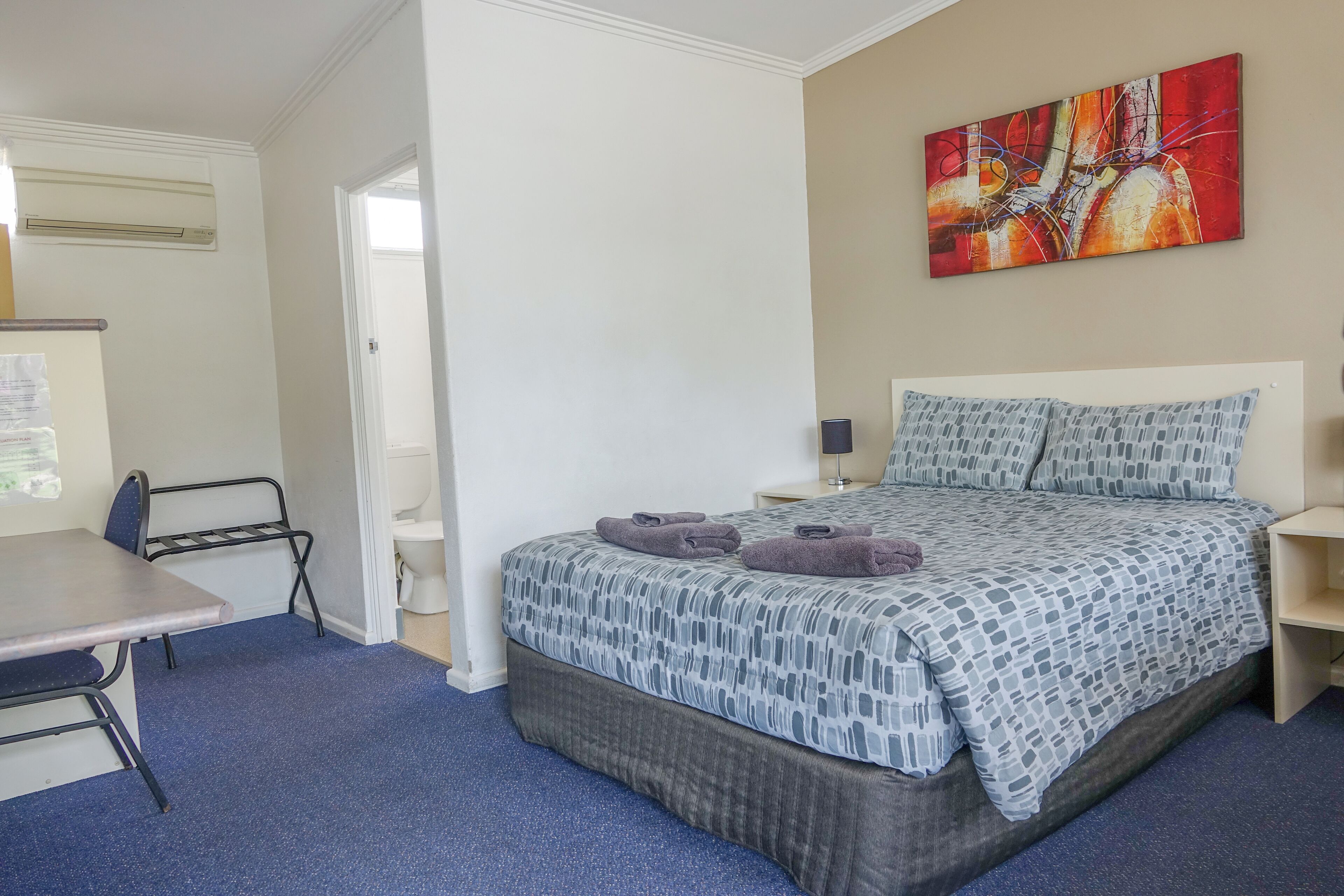 A hotel room with a bed, a desk, a chair, a bedside table, a lamp, and a painting on the wall.