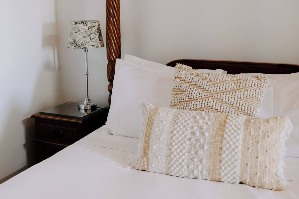 Una cama bien tendida con sábanas blancas y almohadas estampadas, un cabecero de madera y una mesita de noche con una lámpara.