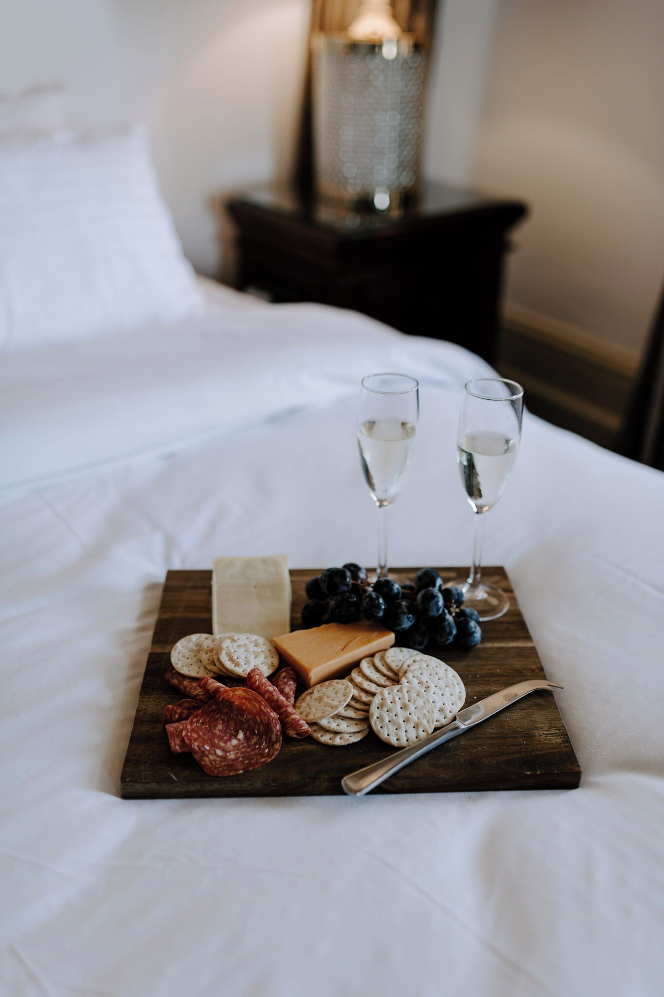 A wooden board with snacks and two glasses of white wine on a bed.