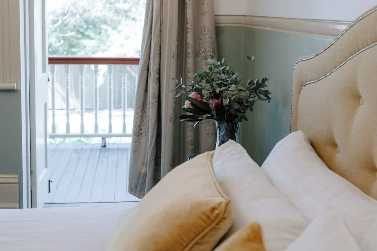 Un dormitorio con cama, cortinas, un jarrón con flores y vistas al balcón.