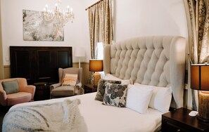 A hotel room with a large bed, a chandelier, a desk with a lamp, a chair, and a large painting on the wall. - The Abbey (Warwick)