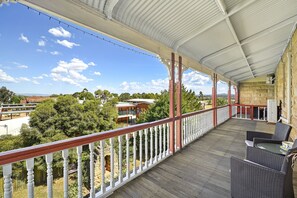 A covered balcony with wicker furniture, a white balustrade, and a view of surrounding buildings and trees.