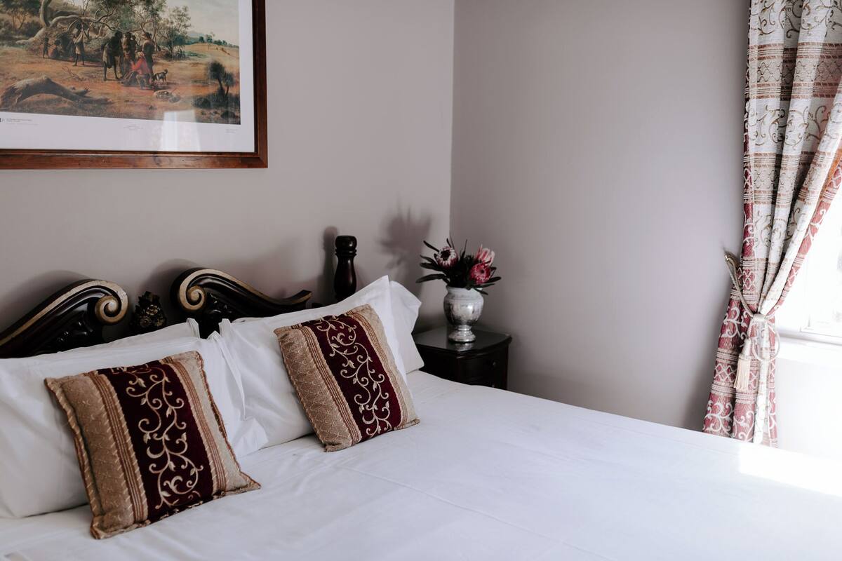 Una cama impecable con sábanas blancas y almohadas decorativas, un cuadro enmarcado en la pared, una mesita con un florero, y una ventana con cortinas.