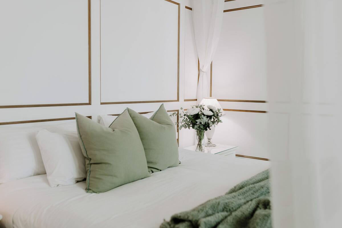 Una cama bien hecha con almohadas blancas y verdes, un jarrón con flores y una mesita de noche con una lámpara.