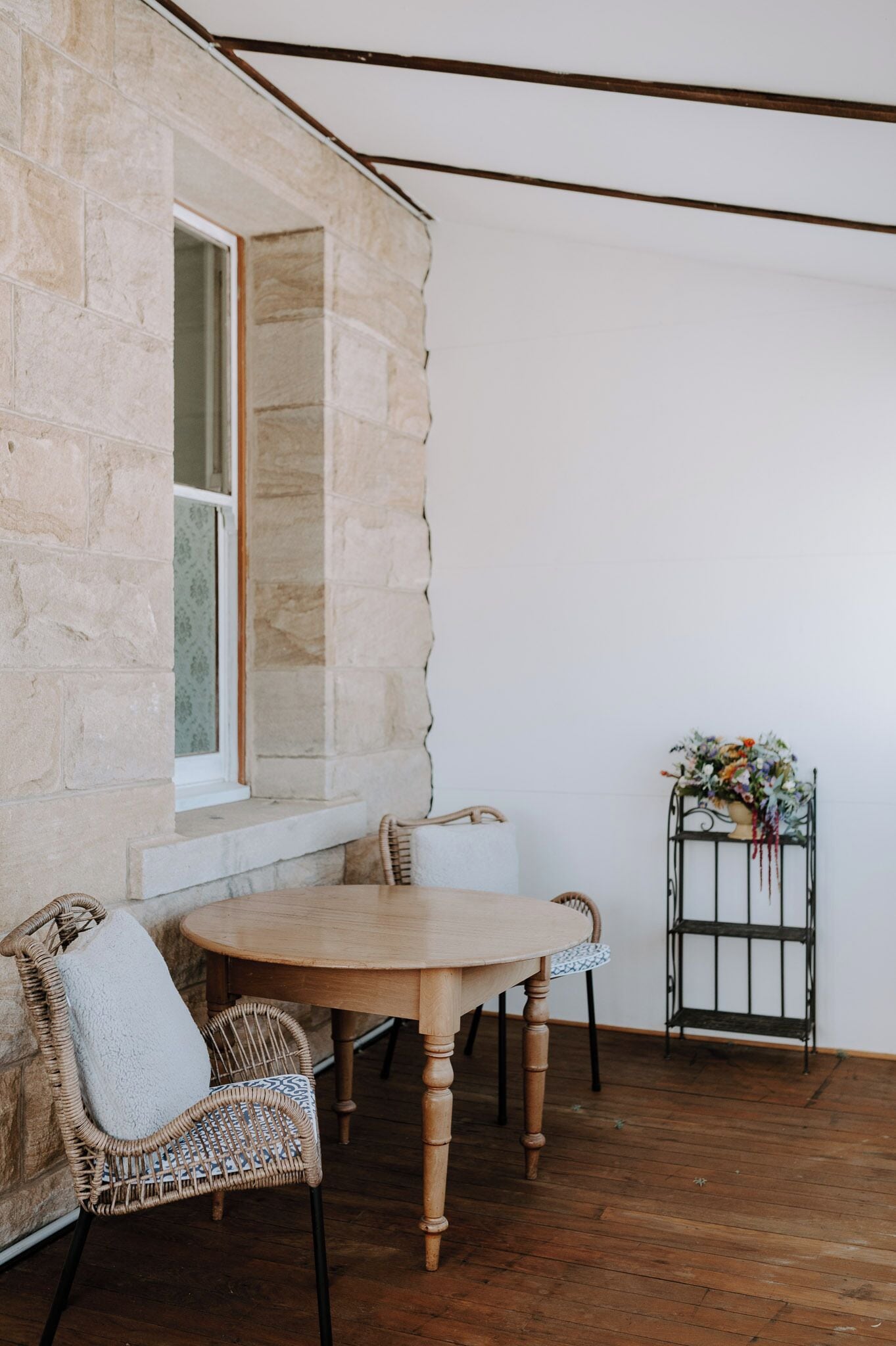 A stone-walled room with a wooden table and chairs, a window, and a metal stand with flowers.