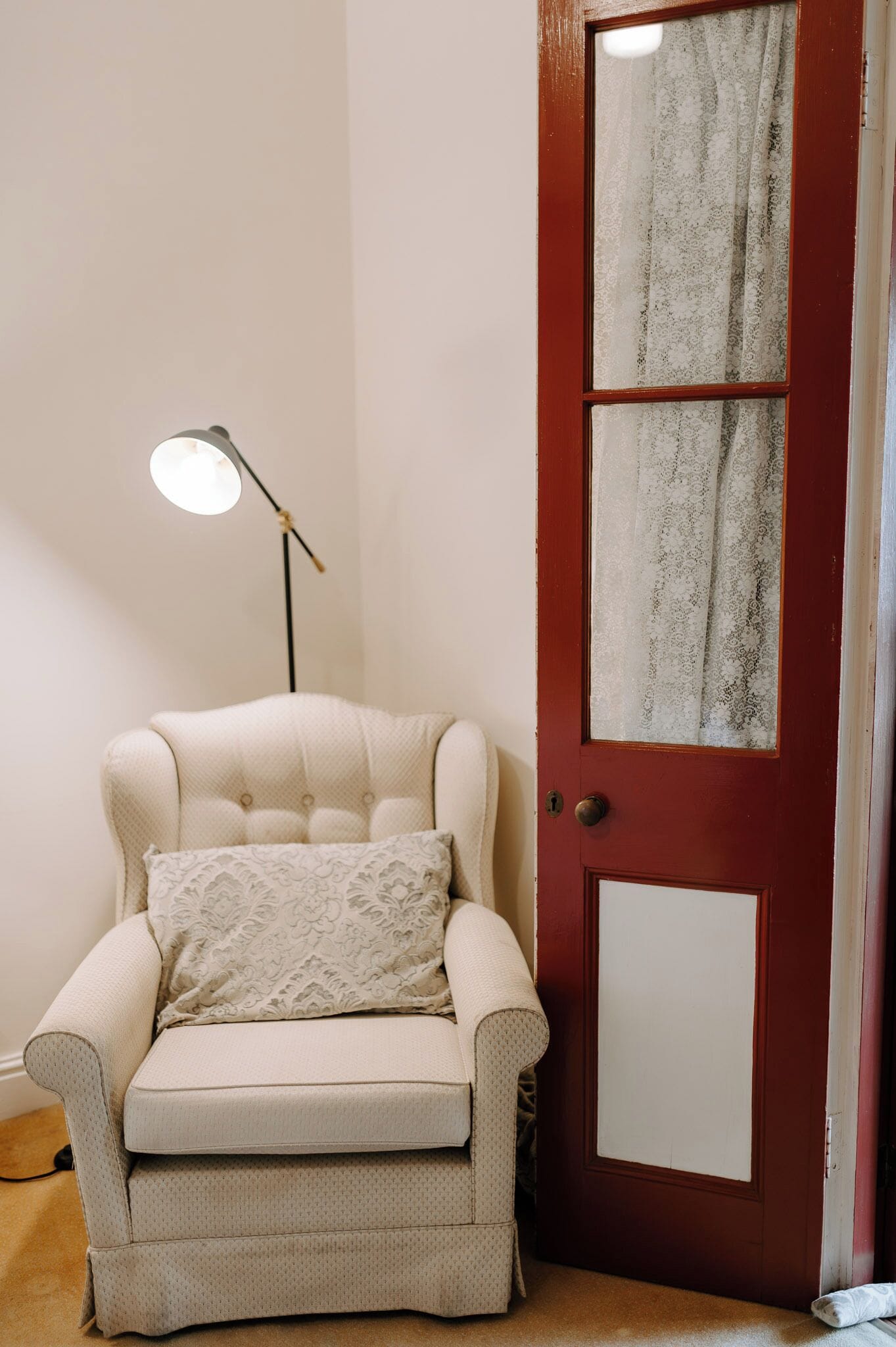 A room with a cream-colored armchair, a floor lamp, and a red door with a frosted glass panel.