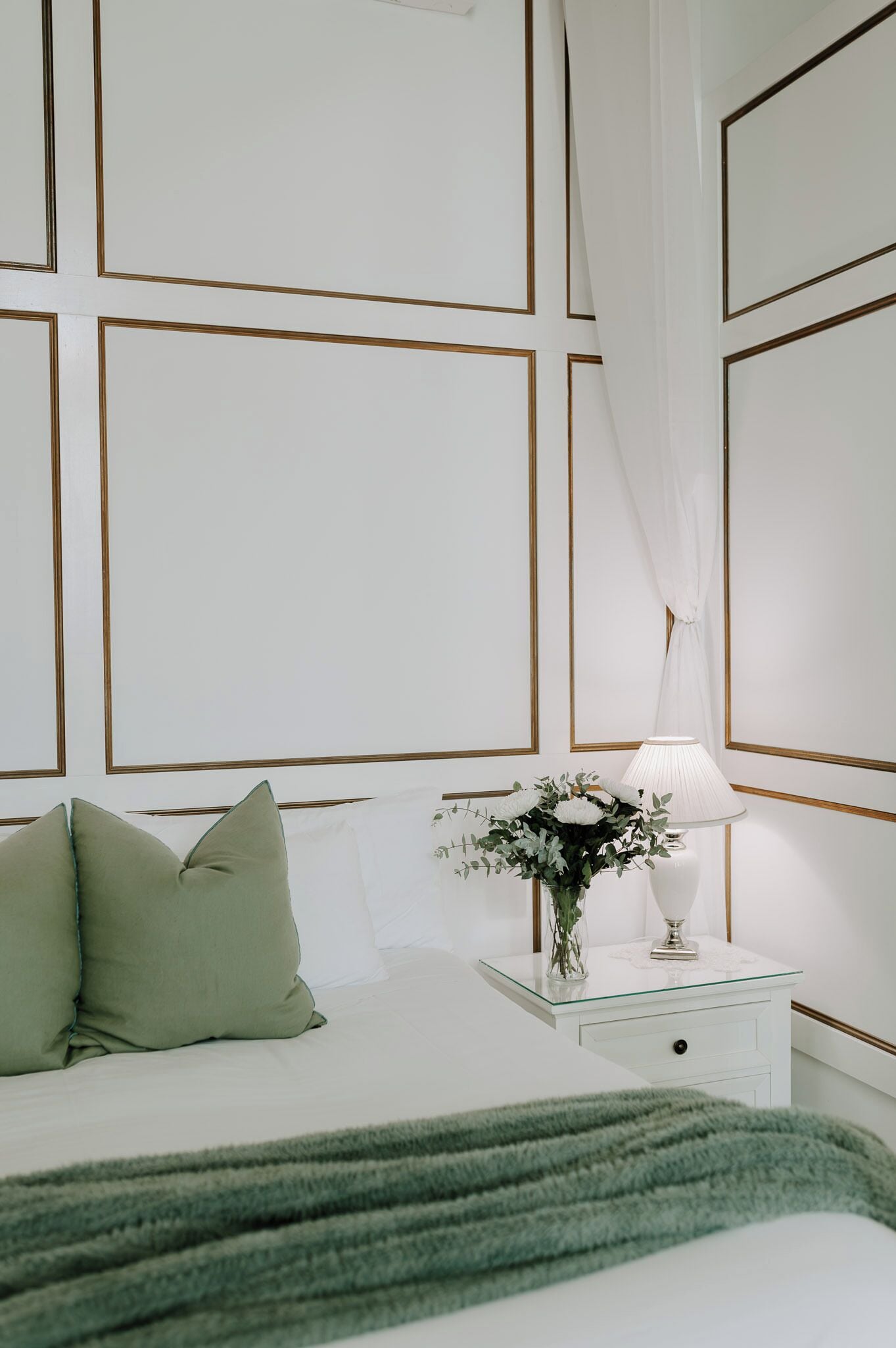 Un dormitorio ordenado con cama, almohadas, una mesita de noche con lámpara y un florero, y una colcha verde.