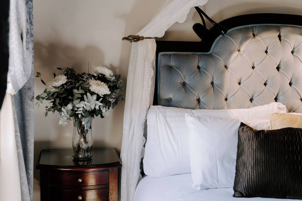 Un dormitorio con cabecera capitonada, una mesita de noche con un jarrón de flores, y una cama con sábanas blancas y almohada oscura.