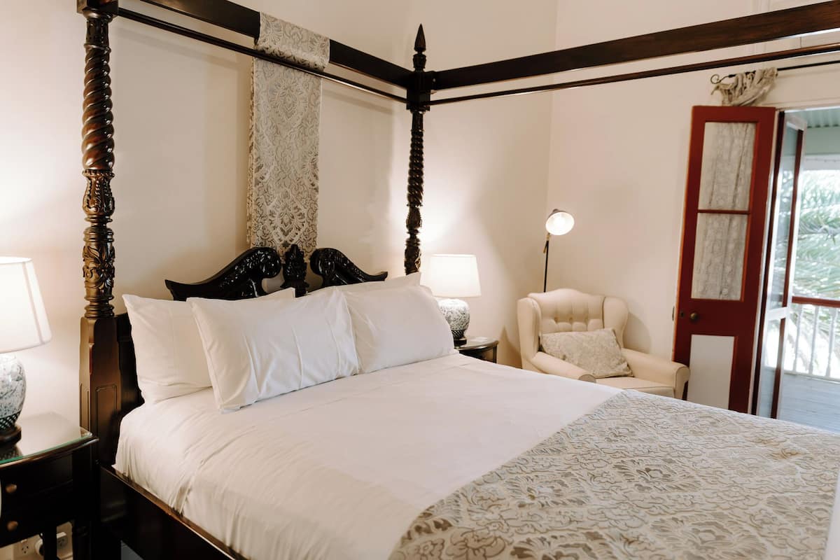 Una cama con dosel, con una colcha blanca y un corredor estampado con motivos florales. Hay un sillón blanco y una lámpara sobre una mesita de noche.