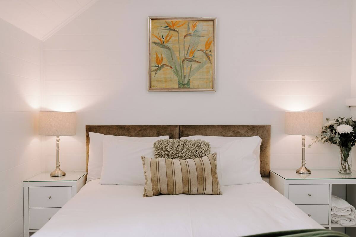 Una cama bien hecha con sábanas blancas, dos lámparas de noche, un cojín decorativo y una obra de arte enmarcada en la pared.