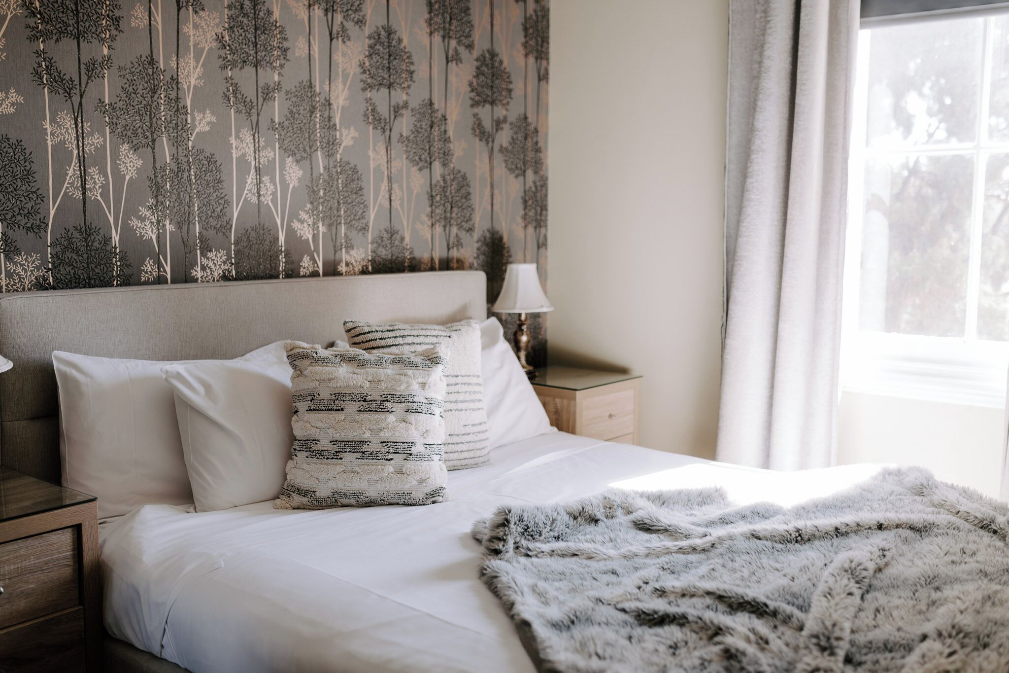 A neatly made bed with white bedding and a grey furry throw blanket. There is a nightstand with a lamp beside the bed. The wall behind the bed has a tree pattern wallpaper.