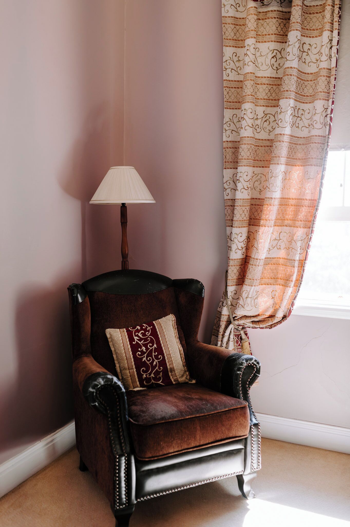 A corner room with a dark brown leather armchair, a white lamp, and a patterned curtain.