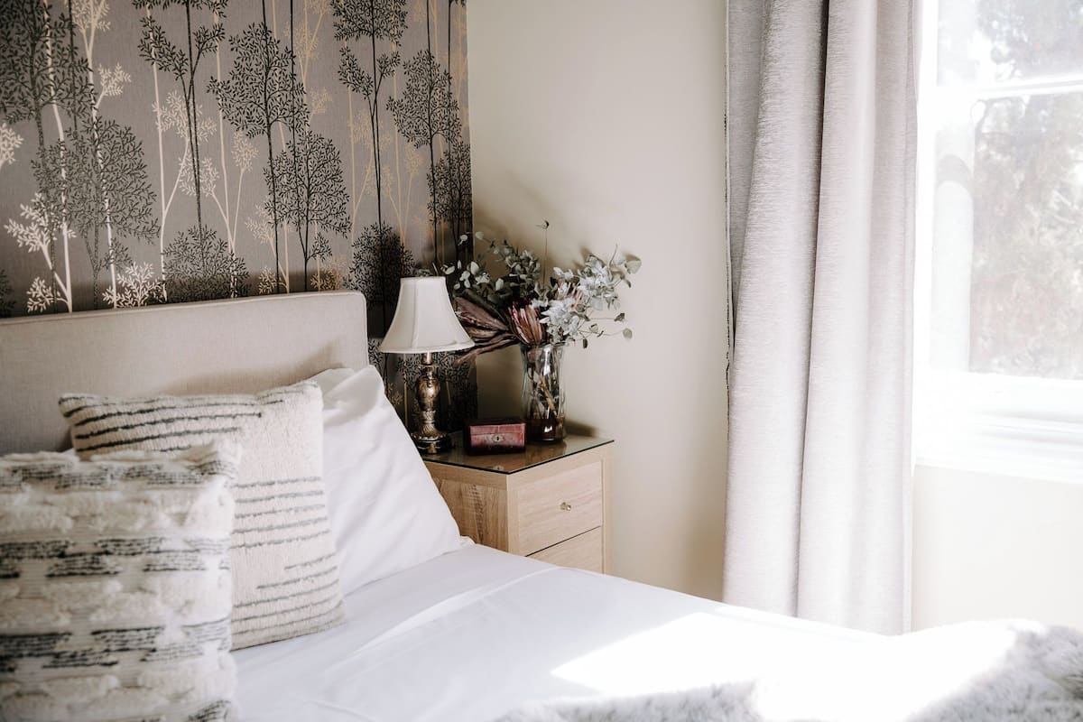 Una cama bien hecha con sábanas blancas y almohadas a rayas, una mesita de noche con una lámpara y un florero, y una pared con un papel tapiz de temática natural.
