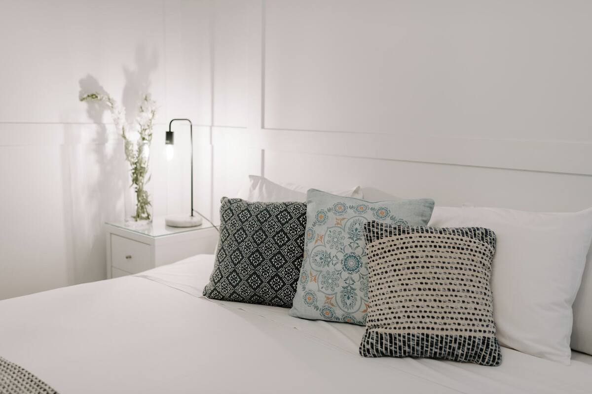 Una cama bien hecha con sábanas blancas y tres almohadas estampadas. Se ve una mesita de noche con una lámpara y un florero con flores.