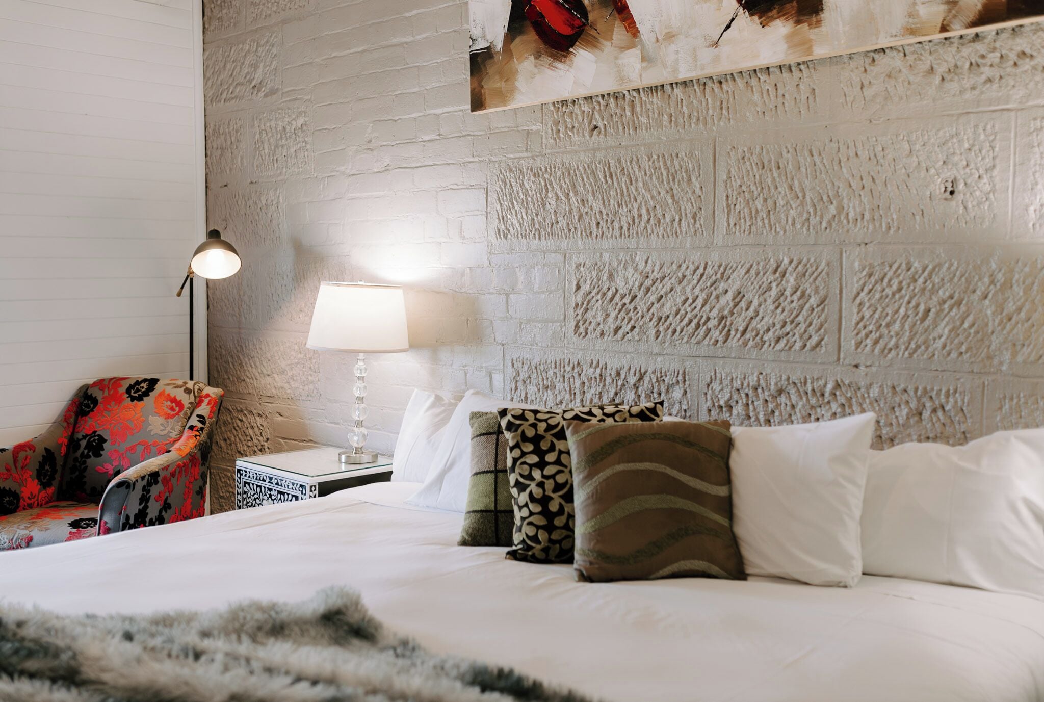 Una habitación de hotel con pared de ladrillo blanco, una cama con almohadas blancas y estampadas, un sillón rojo con motivos florales, una mesita de noche con lámpara y una pequeña mesa con una caja decorativa.