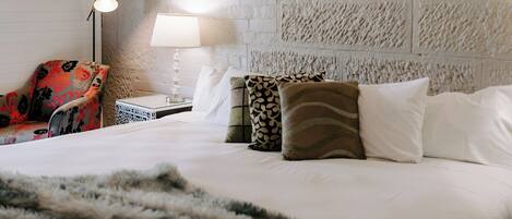 Una habitación de hotel con pared de ladrillo blanco, una cama con almohadas blancas y estampadas, un sillón rojo con motivos florales, una mesita de noche con lámpara y una pequeña mesa con una caja decorativa.