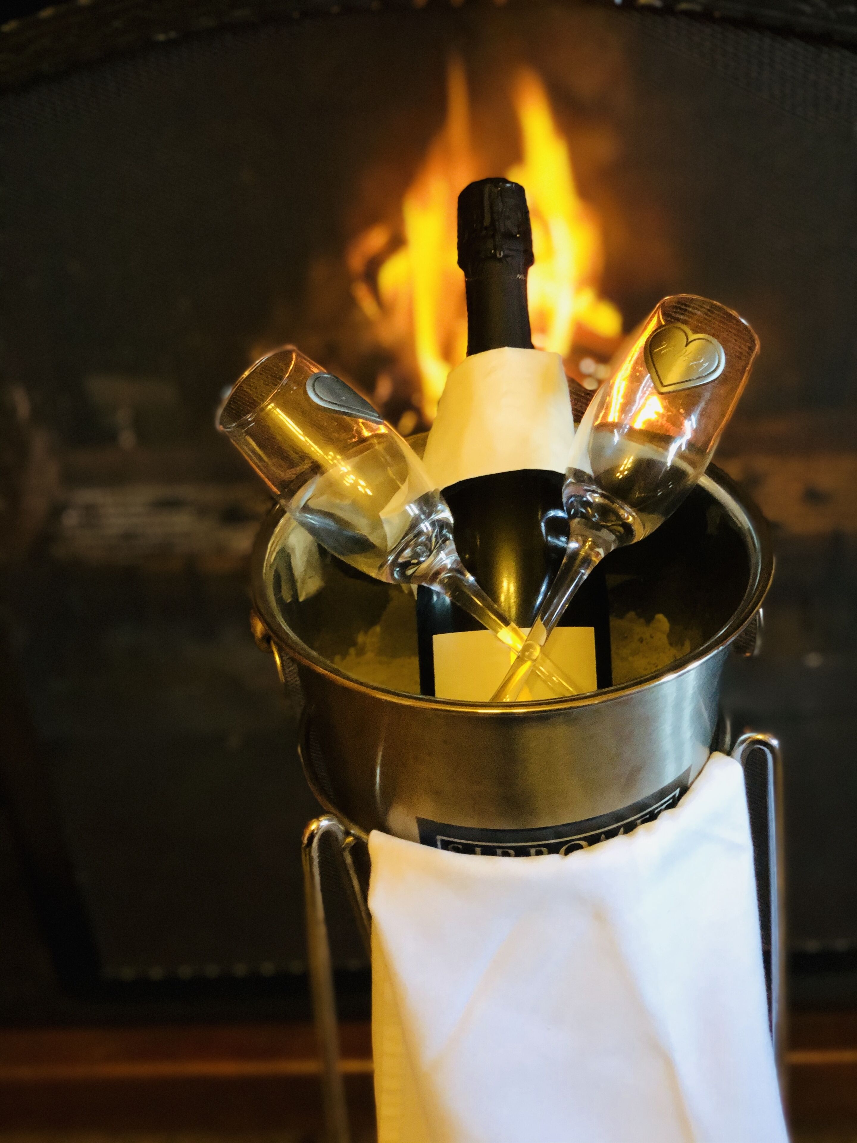 A metal container holding a bottle and glasses, with a lit fire in the background.