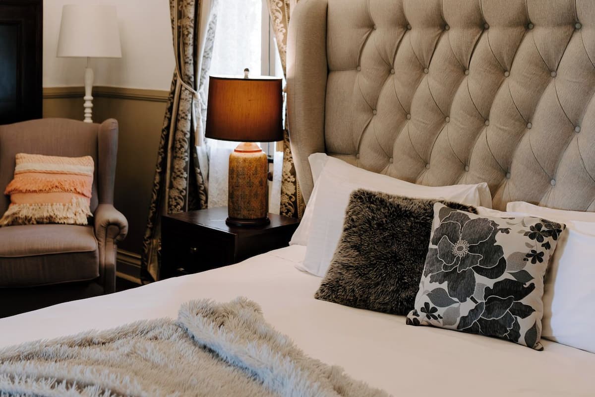 Un dormitorio con cabecera capitonada, cama con varios almohadones, alfombra mullida, mesita de noche con lámpara, una silla y una araña.
