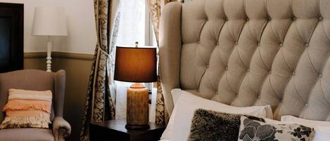 A bedroom with a tufted headboard, a bed with multiple pillows, a fluffy rug, a nightstand with a lamp, a chair, and a chandelier.