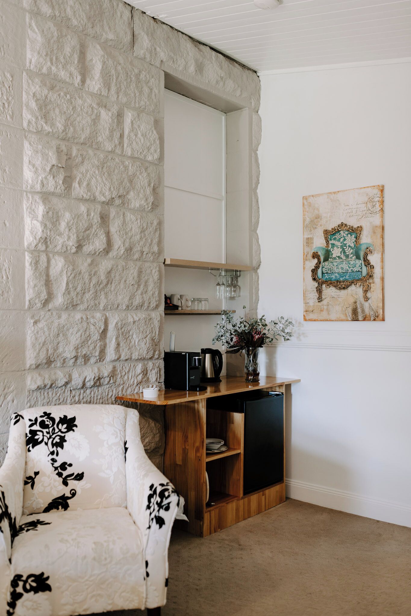 A room with a stone wall, a white armchair, a wooden console table with a vase of flowers, and a framed artwork on the wall.