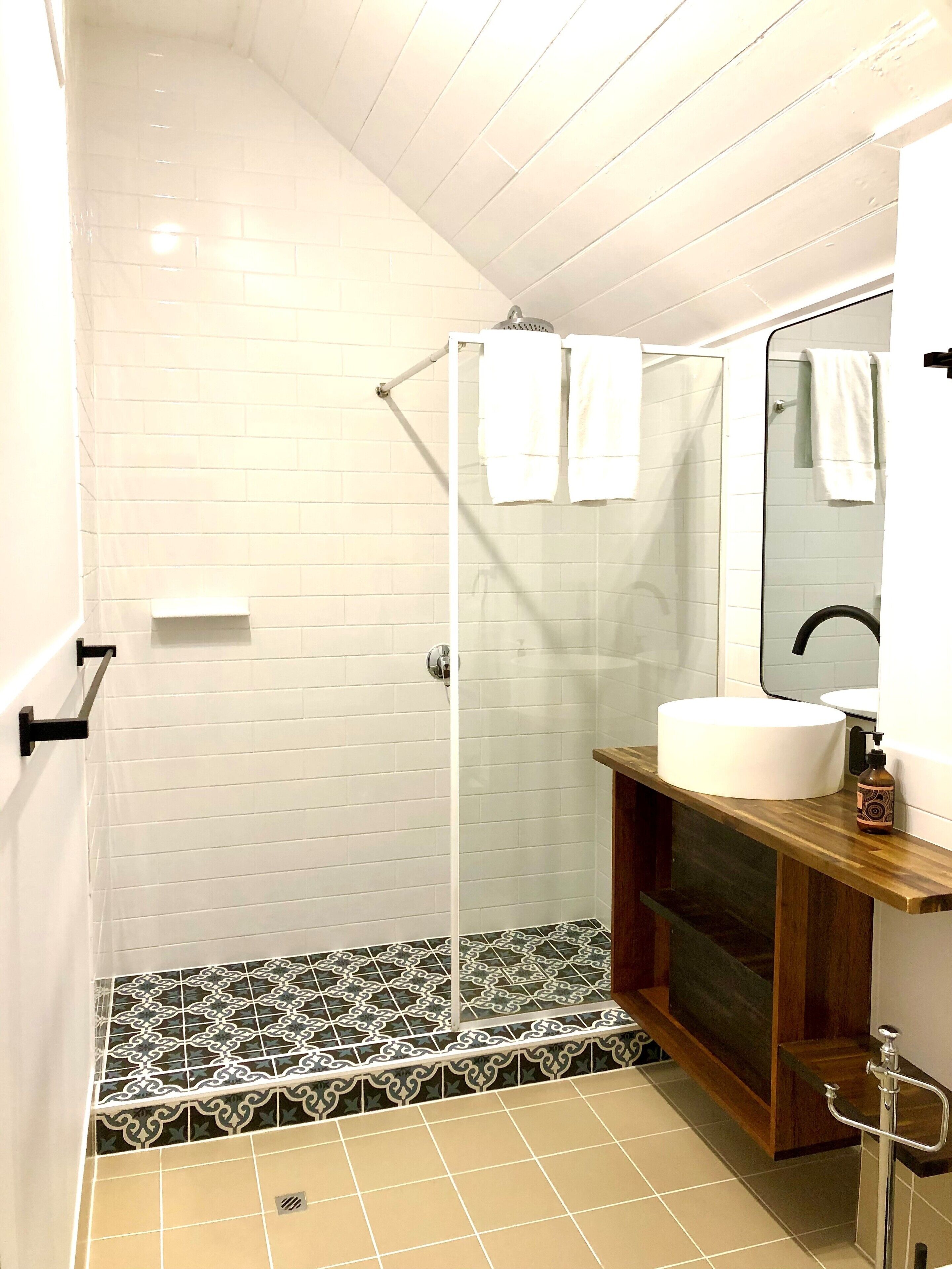 Un baño con ducha, paredes de azulejos blancos, tocador de madera y lavabo.