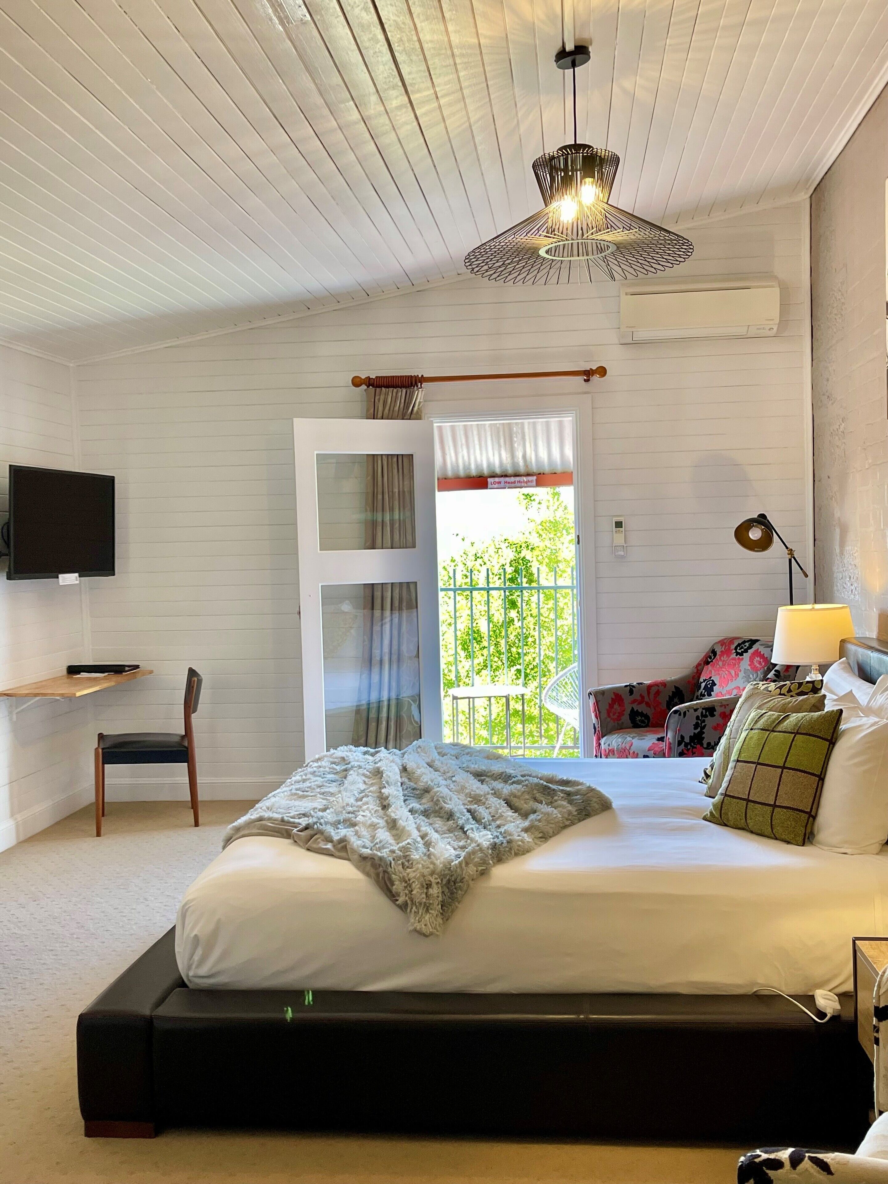 A bedroom with a bed, a chair, a desk, a TV, and a window with a view of greenery.