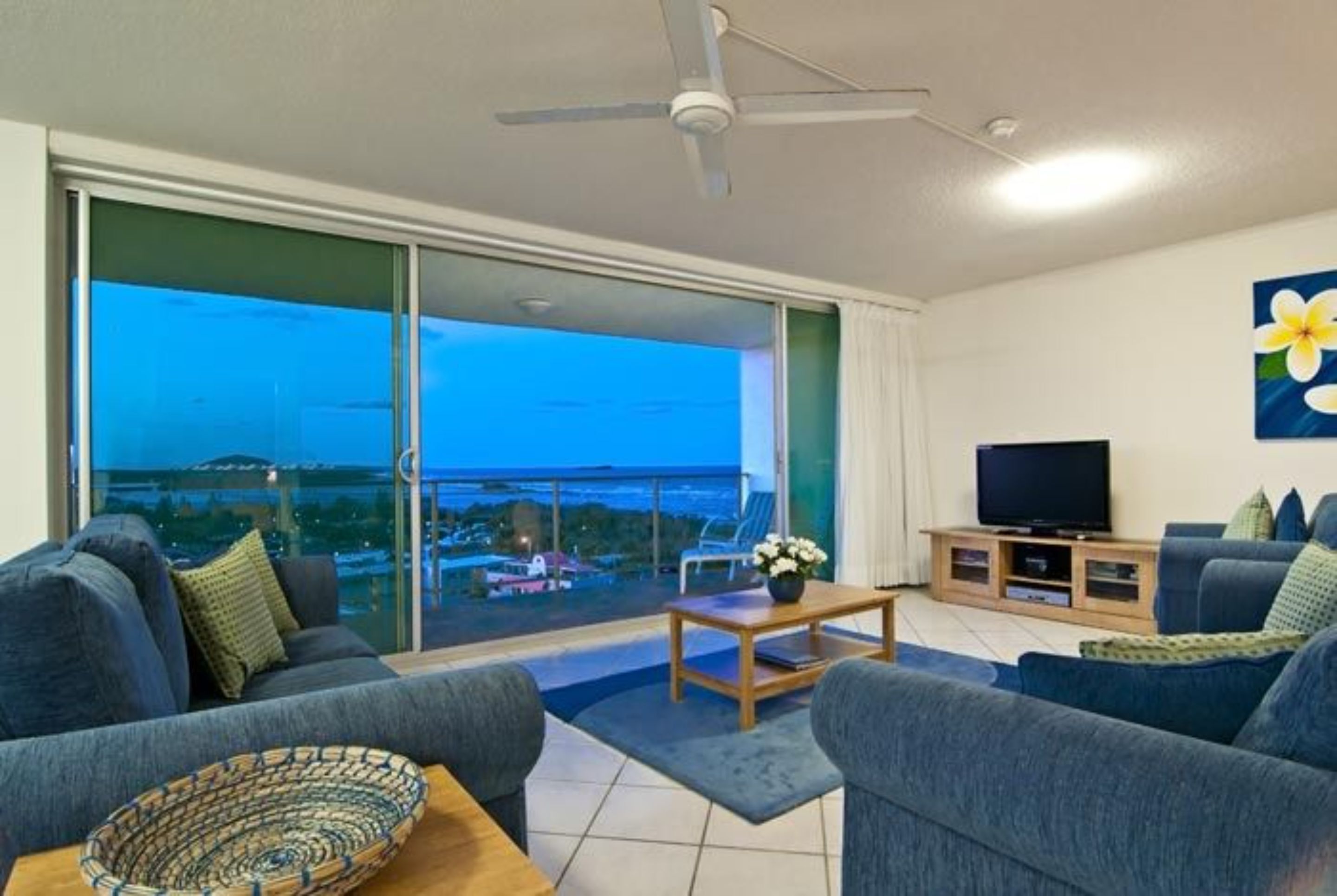 A living room with a sofa, a coffee table, a TV stand, and a view of the city at dusk.