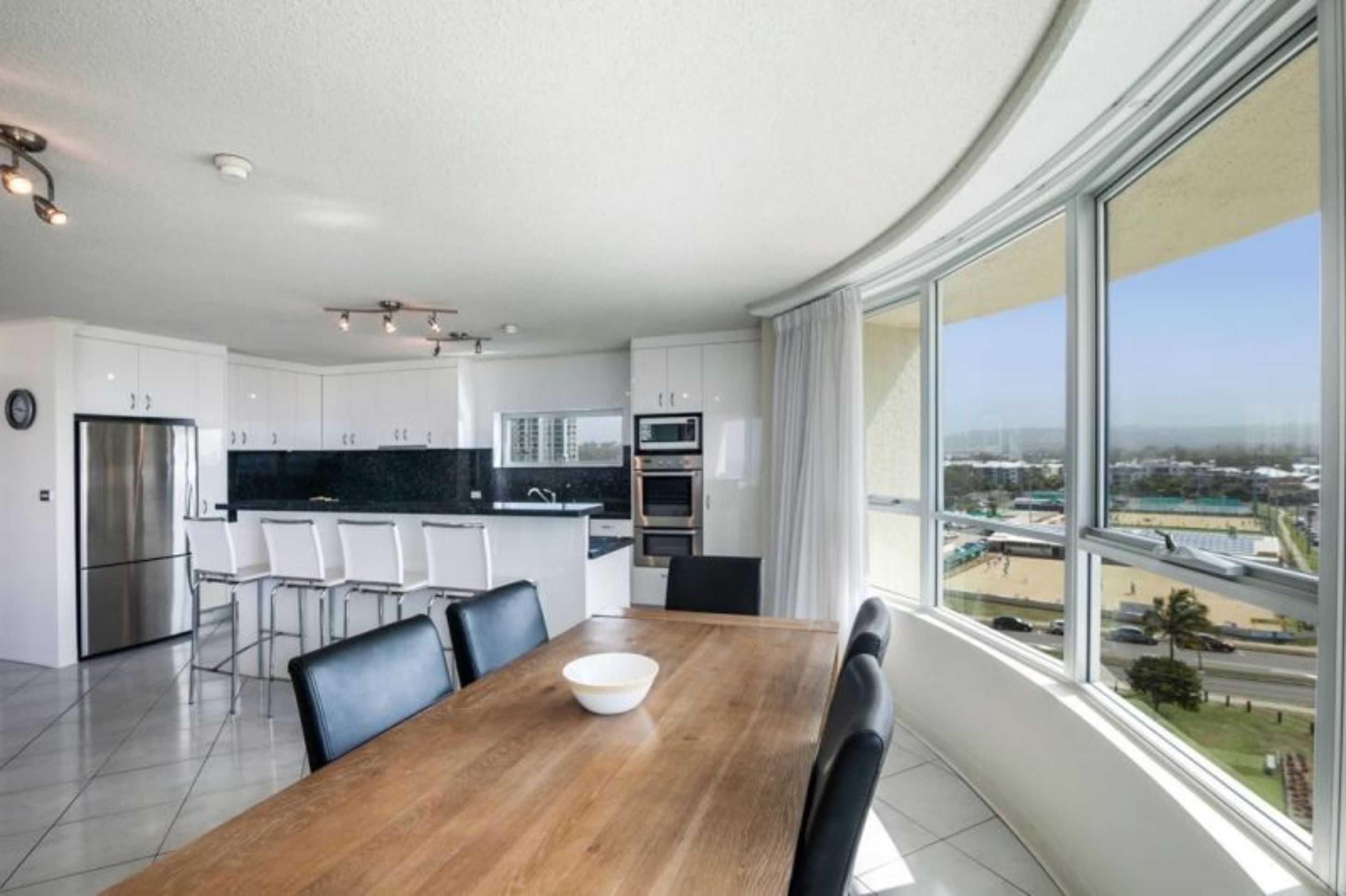 A modern kitchen with white cabinets, a wooden dining table, and a large window offering a city view.