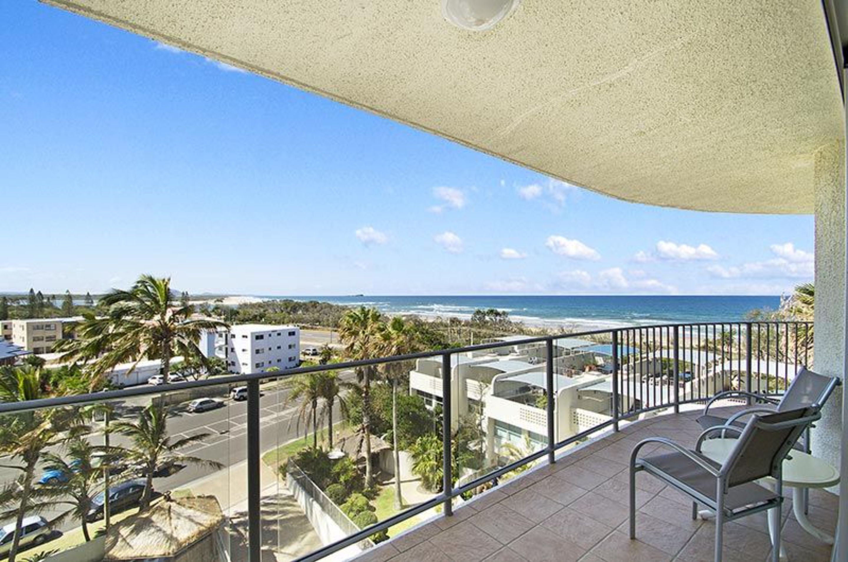 Un balcón con vista al océano y la playa, mobiliario exterior y cielo despejado.