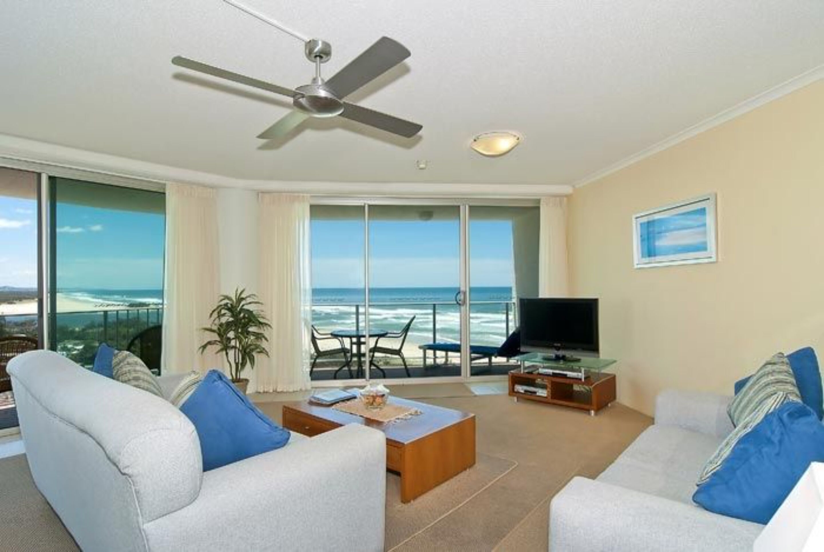A living room with a large window overlooking the ocean, a ceiling fan, and a flat-screen TV on a wooden stand.
