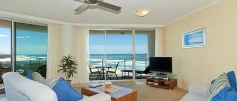 A living room with a large window overlooking the ocean, a ceiling fan, and a flat-screen TV on a wooden stand.
