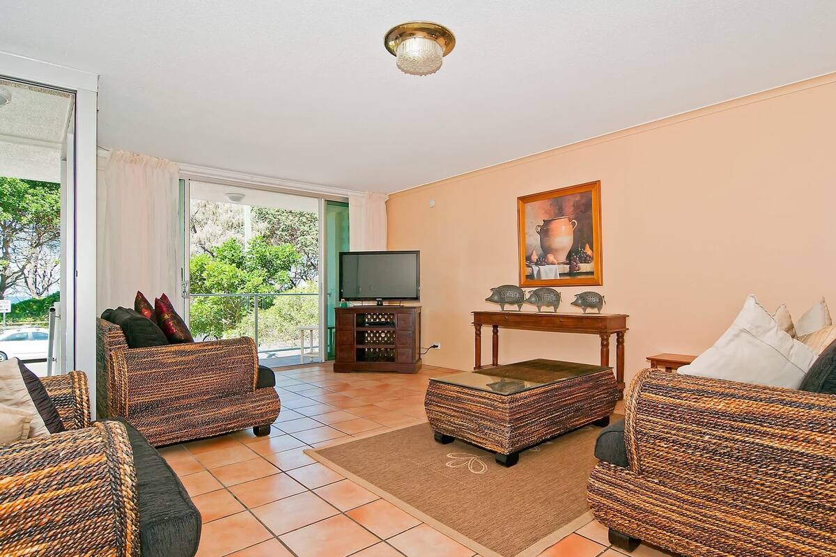 Een woonkamer met rieten meubels, een televisie, een schilderij en uitzicht op groen.