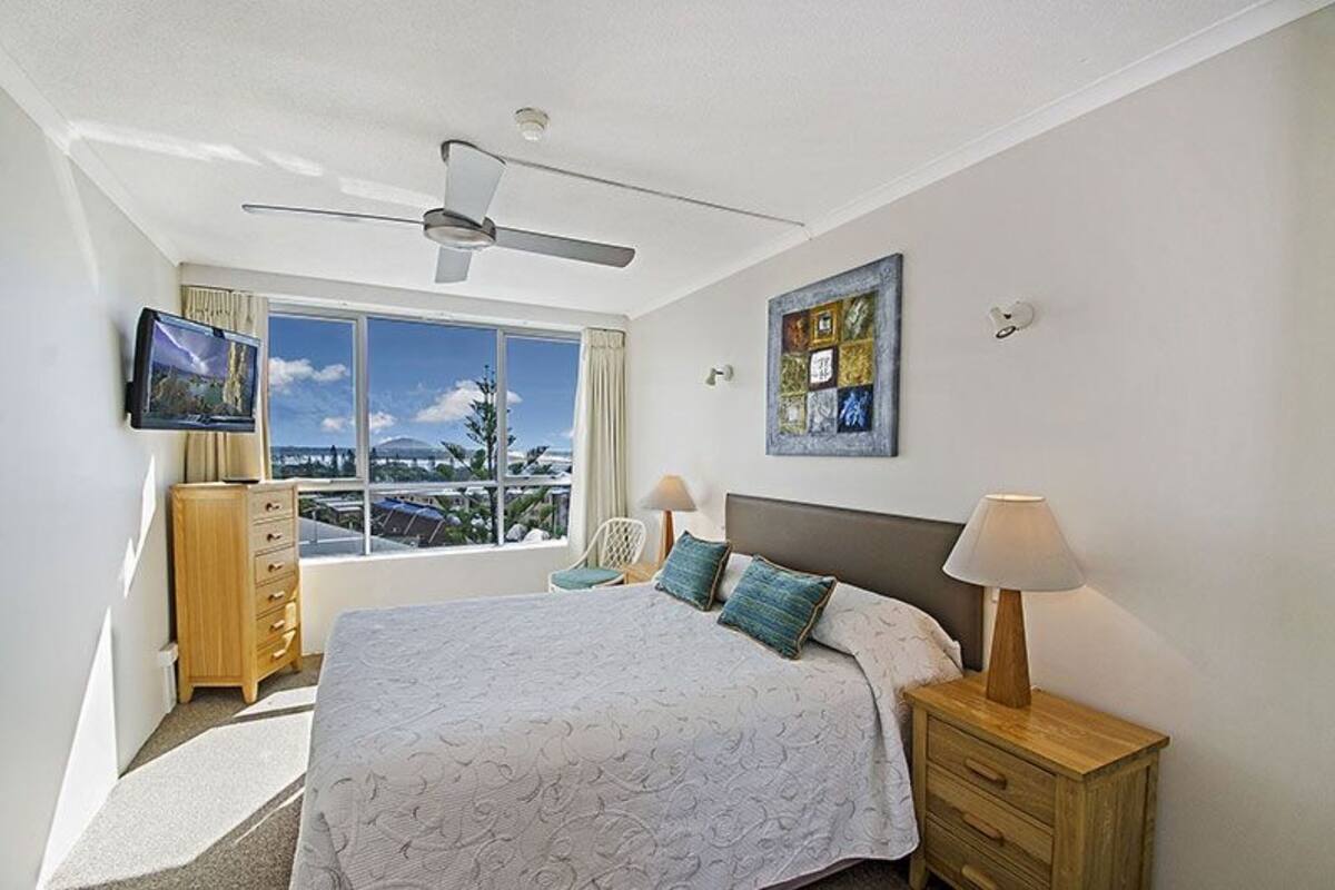 Een slaapkamer met een groot bed, een plafondventilator, een televisie, een raam met uitzicht op de stad en een nachtkastje met een lamp.