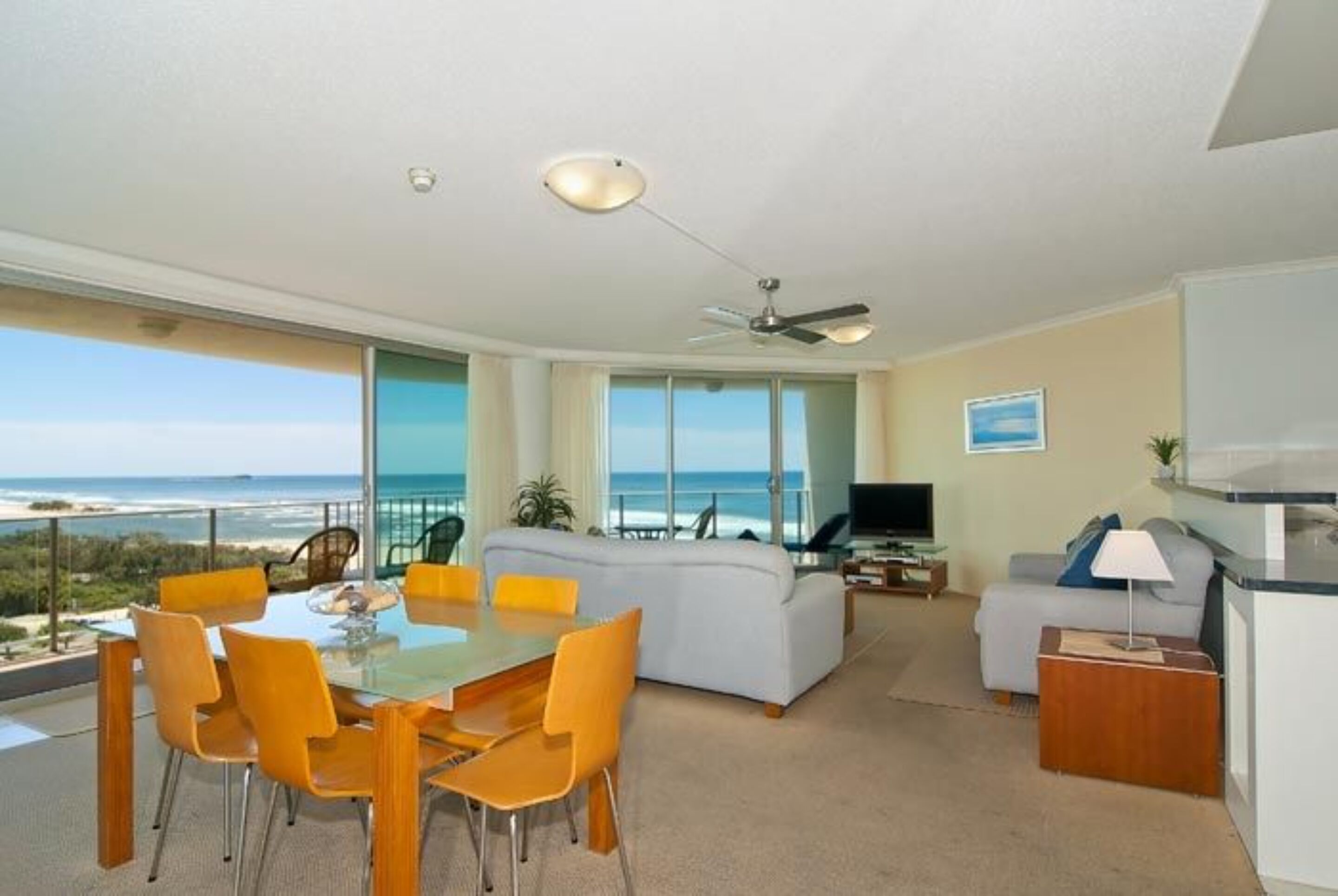 A modern living room with a dining area, a sofa, and a TV. The room has large windows offering a view of the ocean.