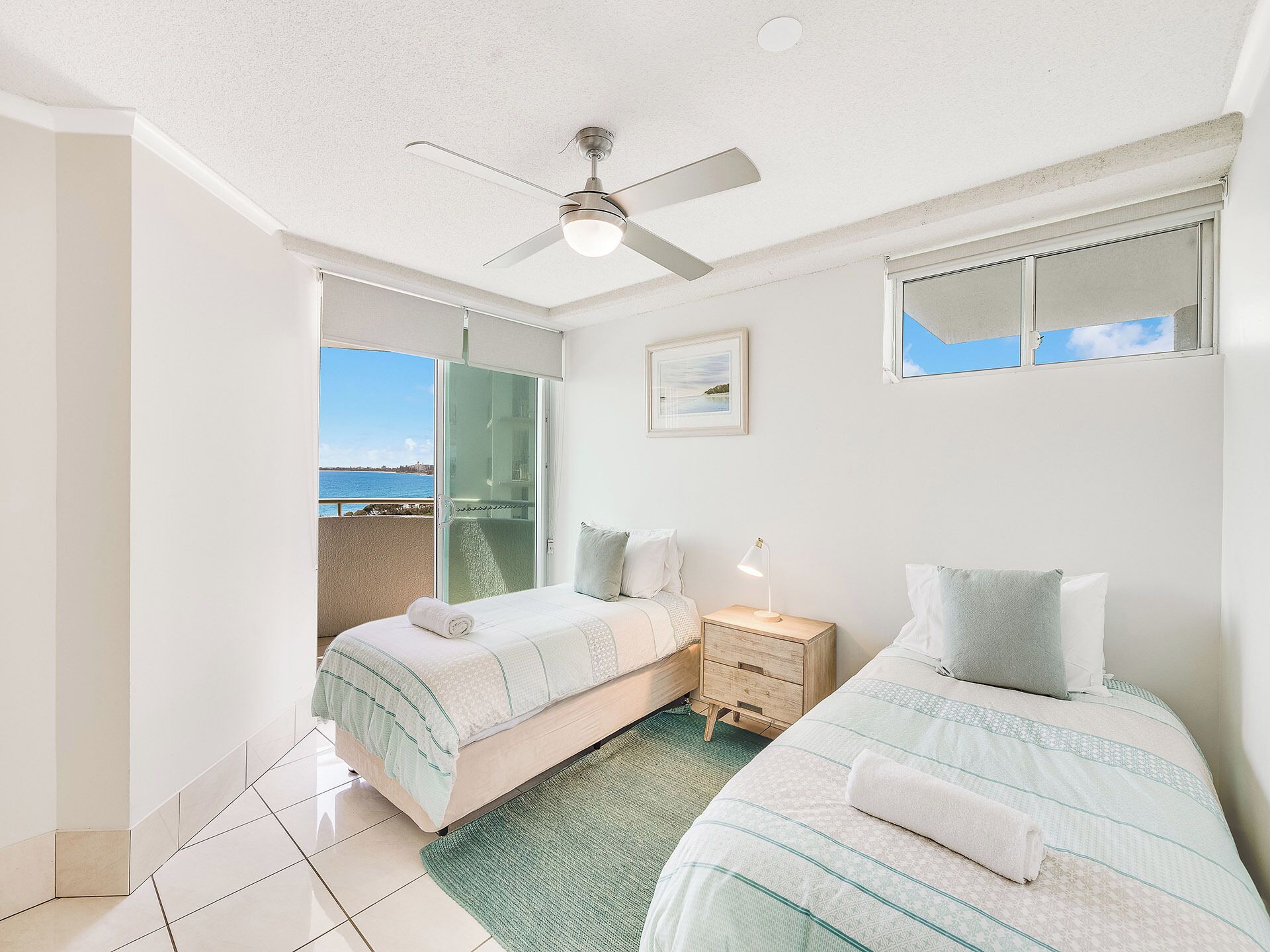 A hotel room with two beds, a ceiling fan, a nightstand, and a view of the sea.