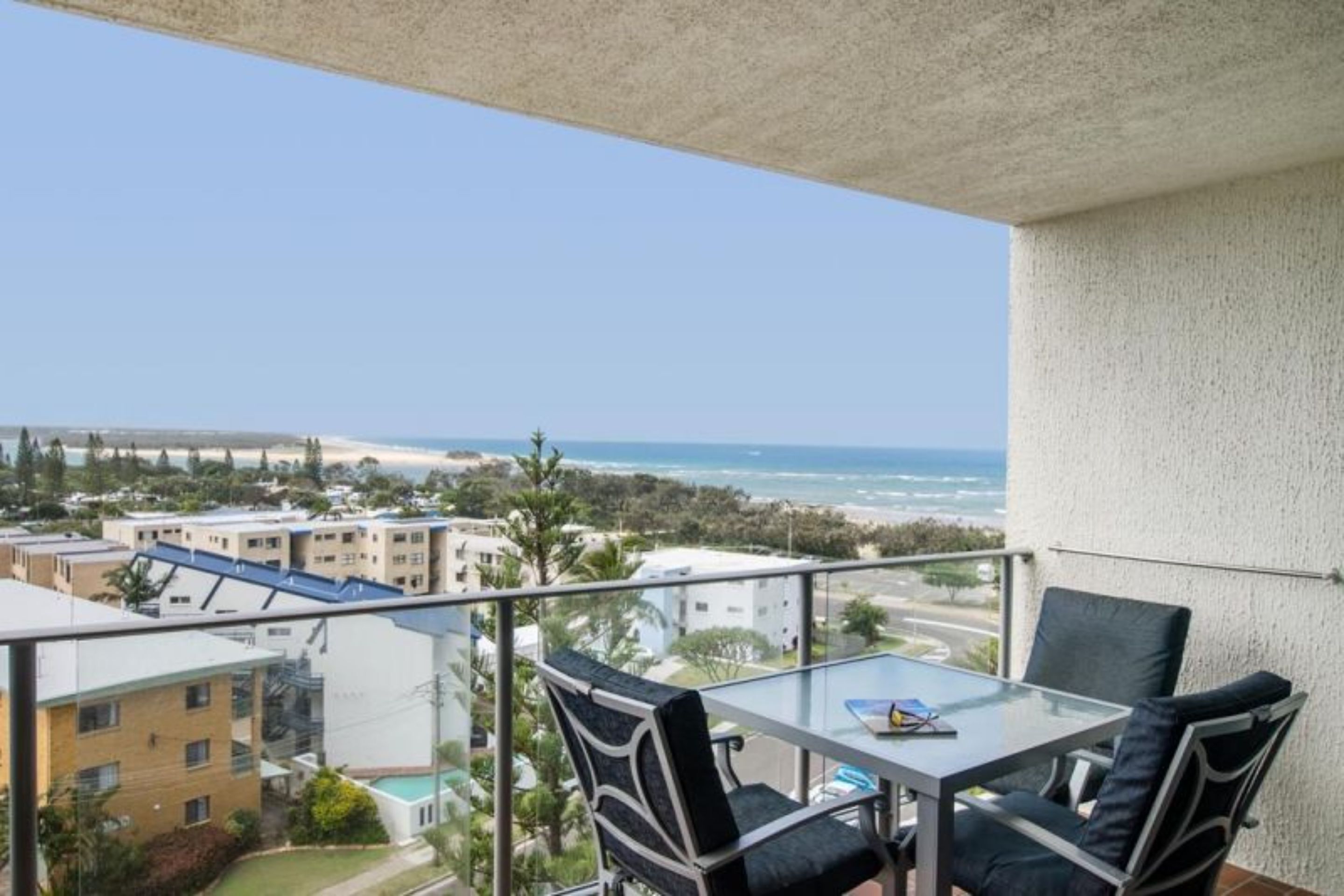 Un balcón con mesa y sillas de vidrio, con vista a una playa y edificios.